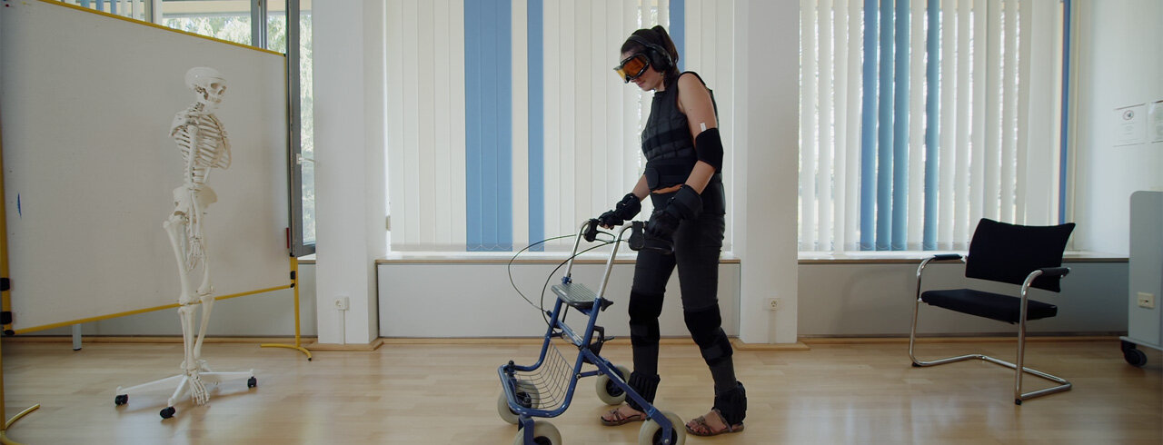 Eine Studierende in der Simulation von Alter. Sie trägt entsprechende Ausrüstung, die das Alter simulieren und stützt sich auf einen Rollator