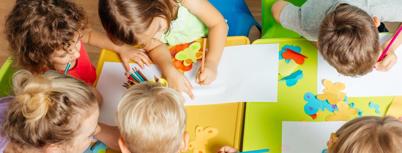 eine Gruppe von Kindern sitzt mit einer Erzieherin am Tisch und malt Bilder