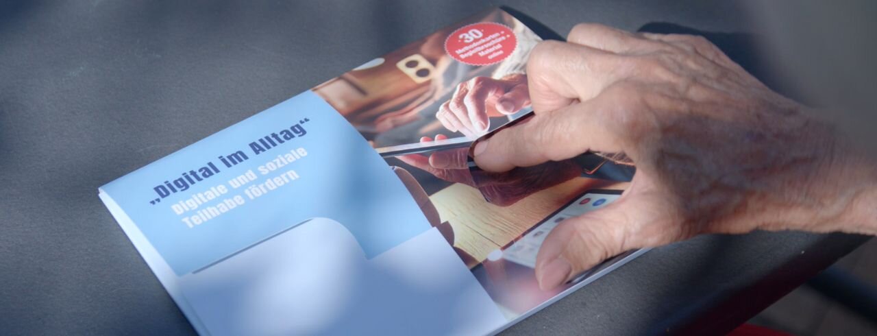 Eine ältere Hand öffnet ein DiBiWohn-Paket, das Programmangebote des DiBiWohn-Projekts zeigt.