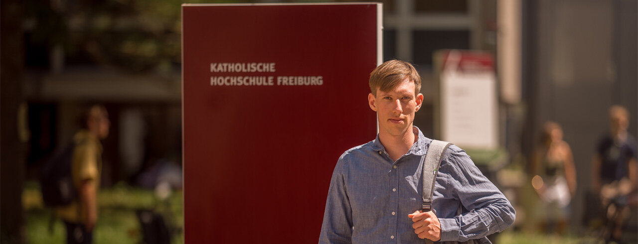 Studierender vor Stehle mit der Aufschrift "Katholische Hochschule Freiburg" vor Campus I