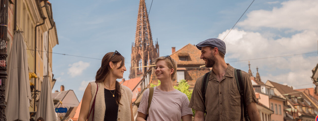 Drei glückliche Studierende der KH Freiburg in der Stadt mit dem Münster im Hintergrund