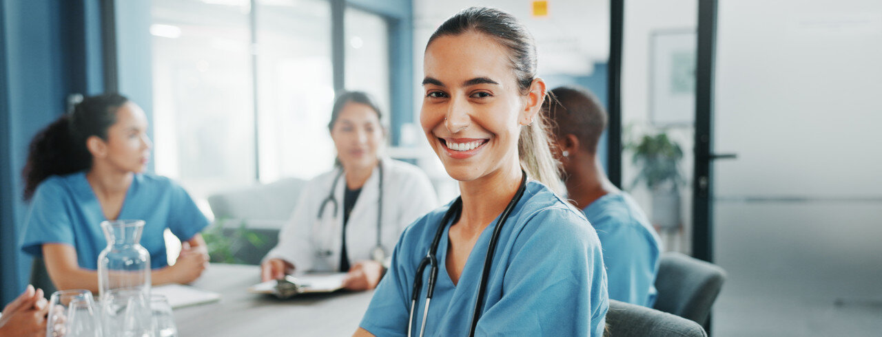 lächelnde Frau am Tisch, hinter ihr eine Gruppe Personen im Gesundheitswesen in einer Besprechung